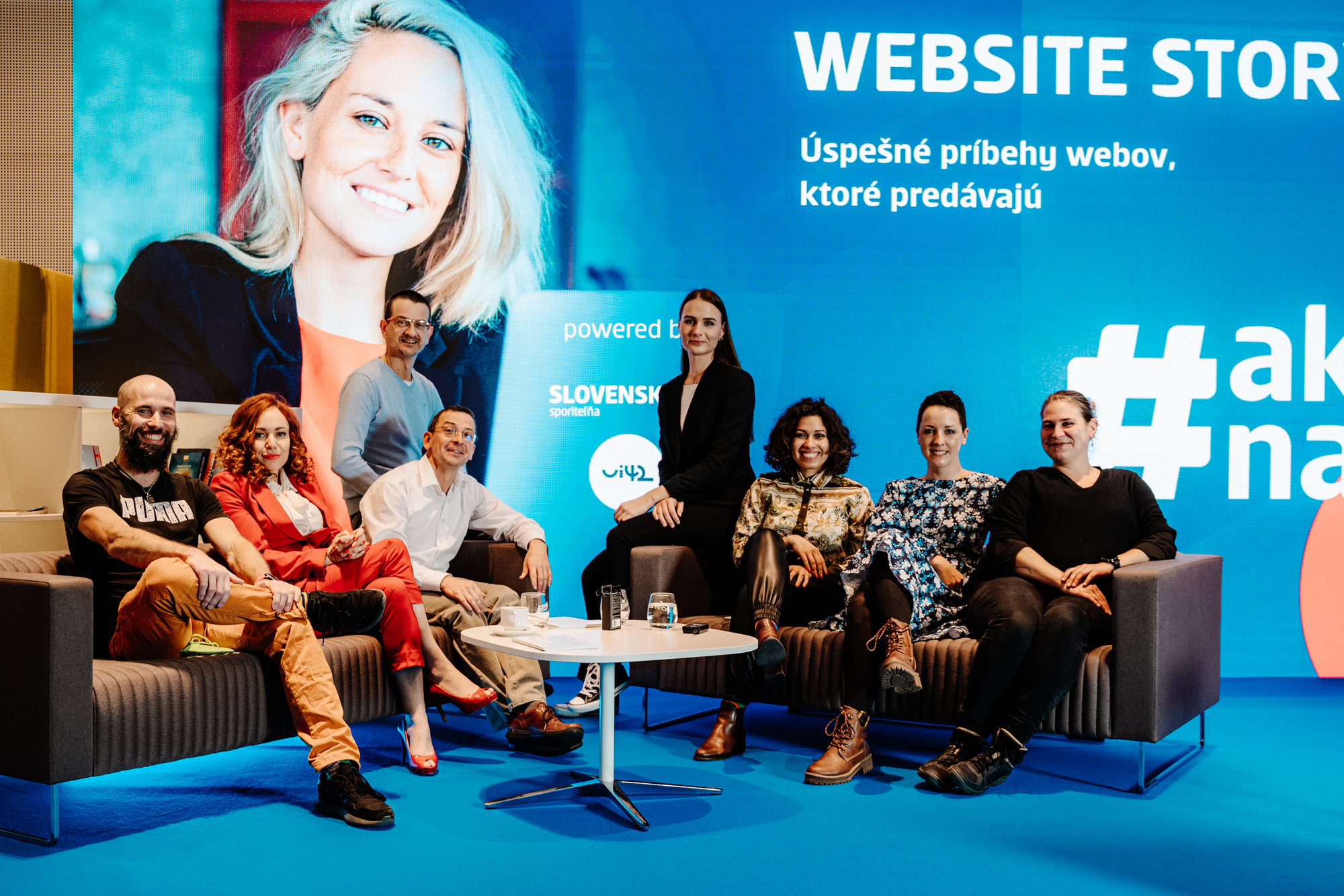 Skupina lidí sedících na pohovkách během akce Website Story, s velkým modrým pozadím a nápisy o úspěšných příbězích webů.