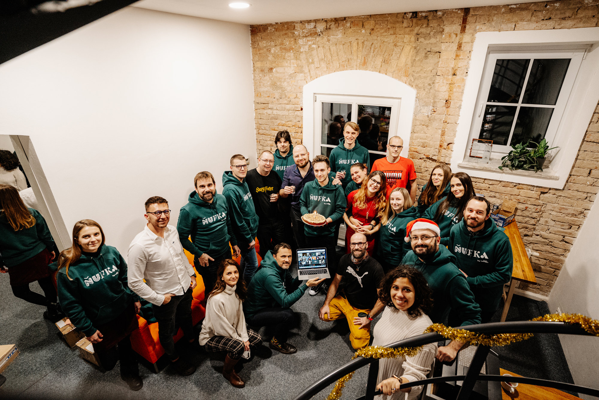 Skupinová fotografie lidí v interiéru s cihlovou stěnou, někteří jsou oblečeni v zelených mikinách s nápisem 'NUFKA', usmívají se a drží dort a laptop.