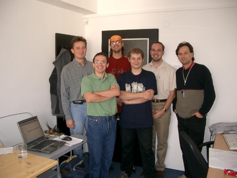 Skupina šesti mužů stojí spolu v kanceláři, usmívají se a jsou neformálně oblečeni. Na stole vedle nich je laptop a sklenice s vodou.