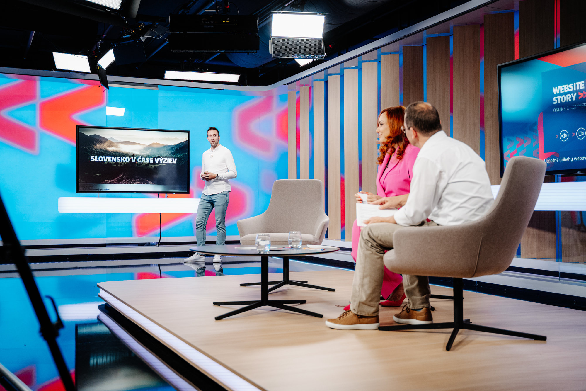 Diskusní studio se třemi lidmi, kde jeden stojí u obrazovky s textem 'Slovensko v čase výzev' a další dva sedí na židlích a sledují ho. Prostředí je moderní s barevnými světly a kamerou v popředí.