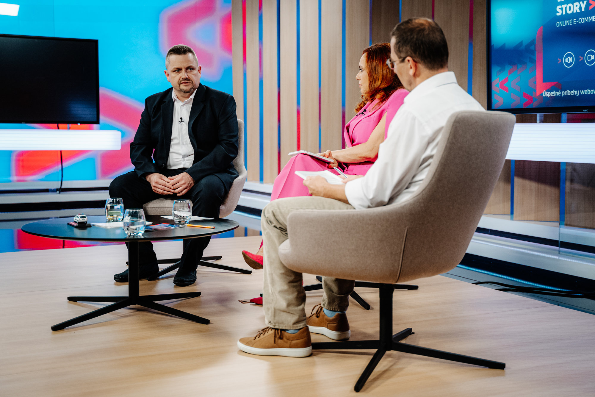Diskusní panel v televizním studiu se třemi lidmi sedícími v křeslech kolem stolku. Moderátor a hosté diskutují na téma přítomnosti nebo budoucnosti.