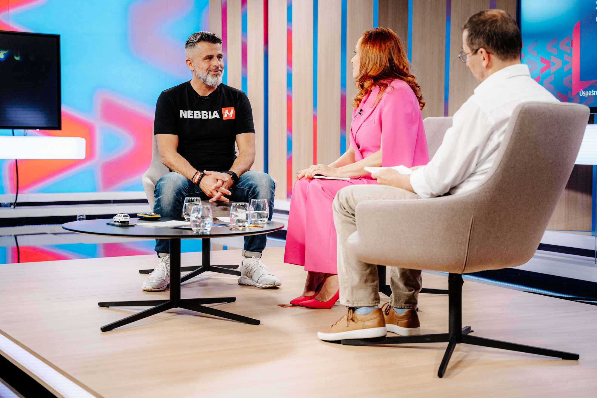 Diskuse tří osob v televizním studiu, muž v tričku Nebbia a žena v růžových šatech