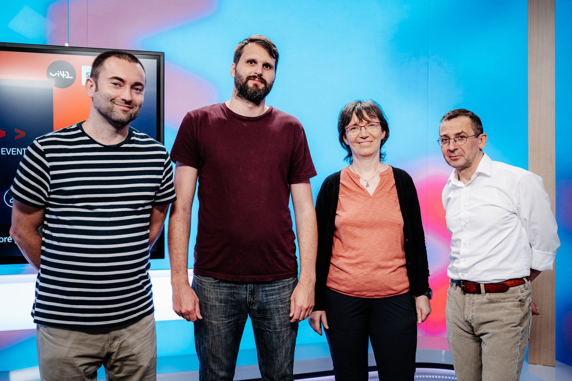 Čtyři lidé stojící před barevným pozadím, dva muži v tričkách, jedna žena v oranžovém tričku a muž v bílé košili.
