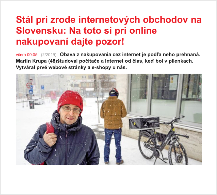 Muž v červené čepici stojí na zasněžené ulici s budovou v pozadí, vedle zaparkovaného kola.