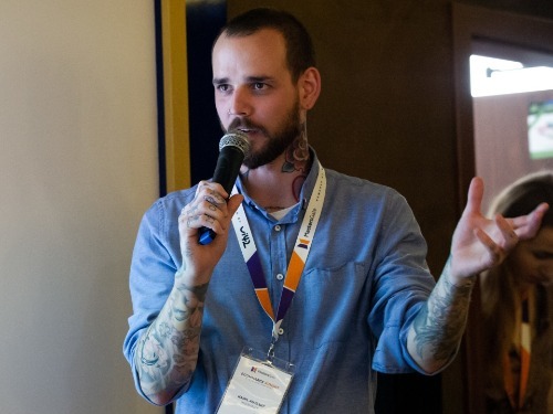 Muž s tetováním na rukou drží mikrofon a mluví na konferenci MastersGate.