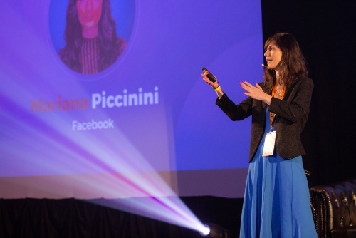 Žena prezentující na konferenci MastersGate s mikrofonem a dálkovým ovladačem v ruce, oblečená v modrých šatech a saku.