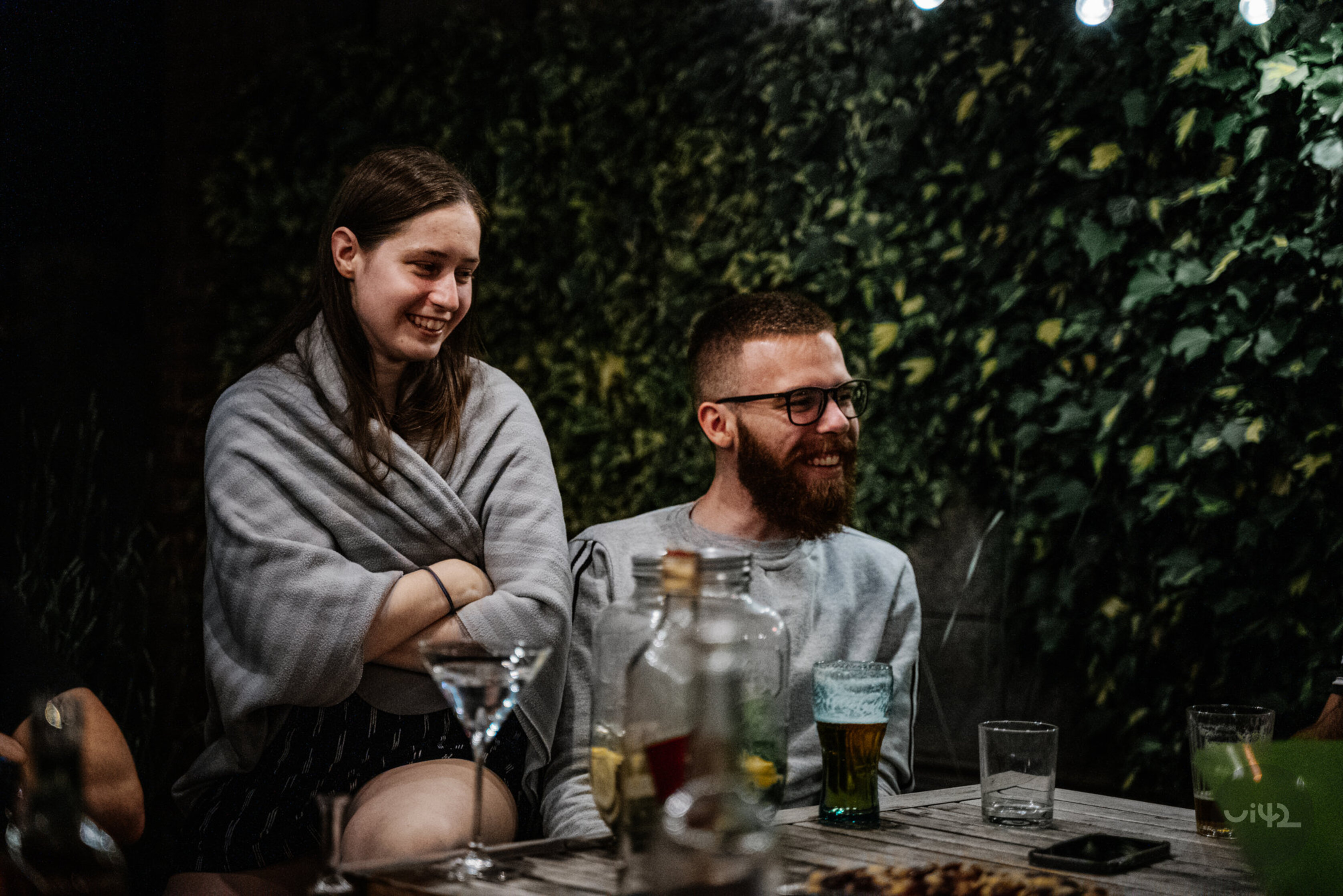 Dva lidé sedí venku u stolu, povídají si a smějí se. Na stole jsou sklenice s nápoji a v pozadí je zelená stěna rostlin.