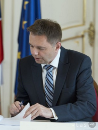 Muž v obleku podepisuje dokument u stolu s vlajkami v pozadí.