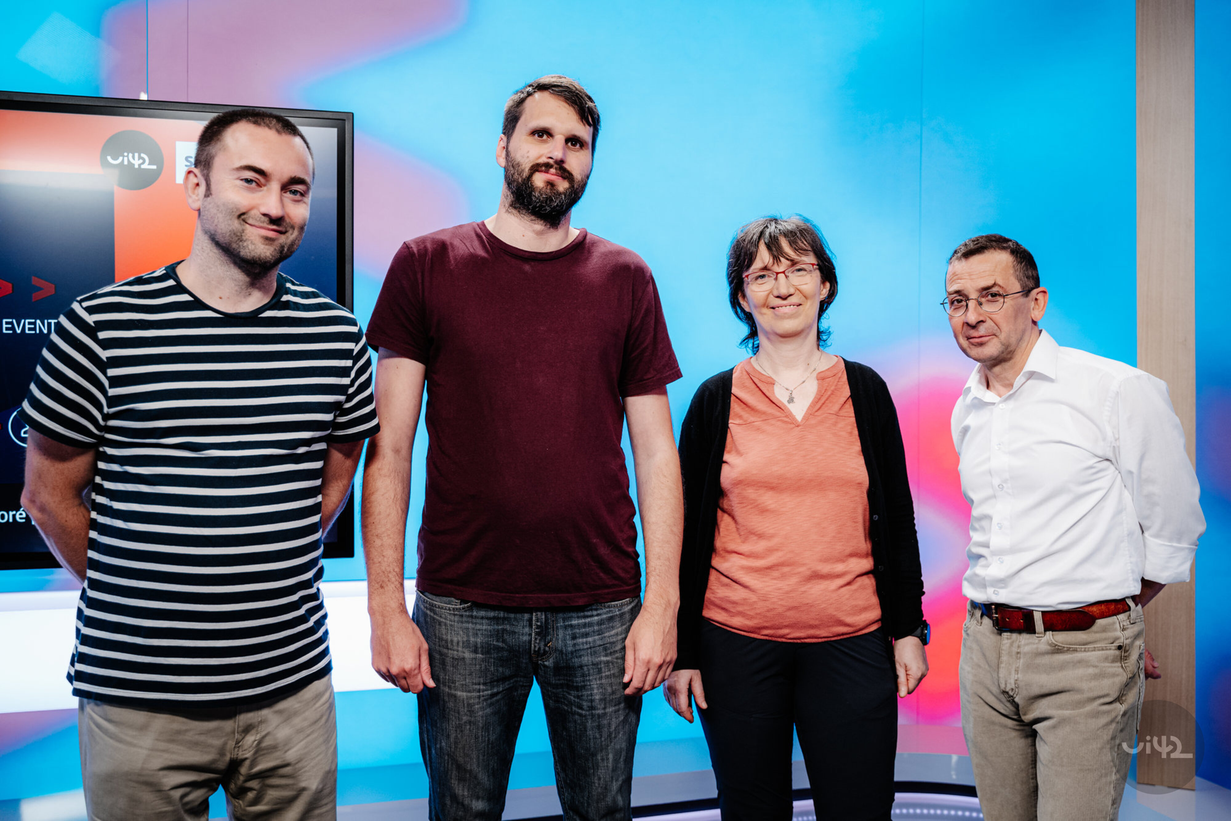 Čtyři lidé stojící před barevným pozadím, dva muži v tričkách, jedna žena v oranžovém tričku a muž v bílé košili.