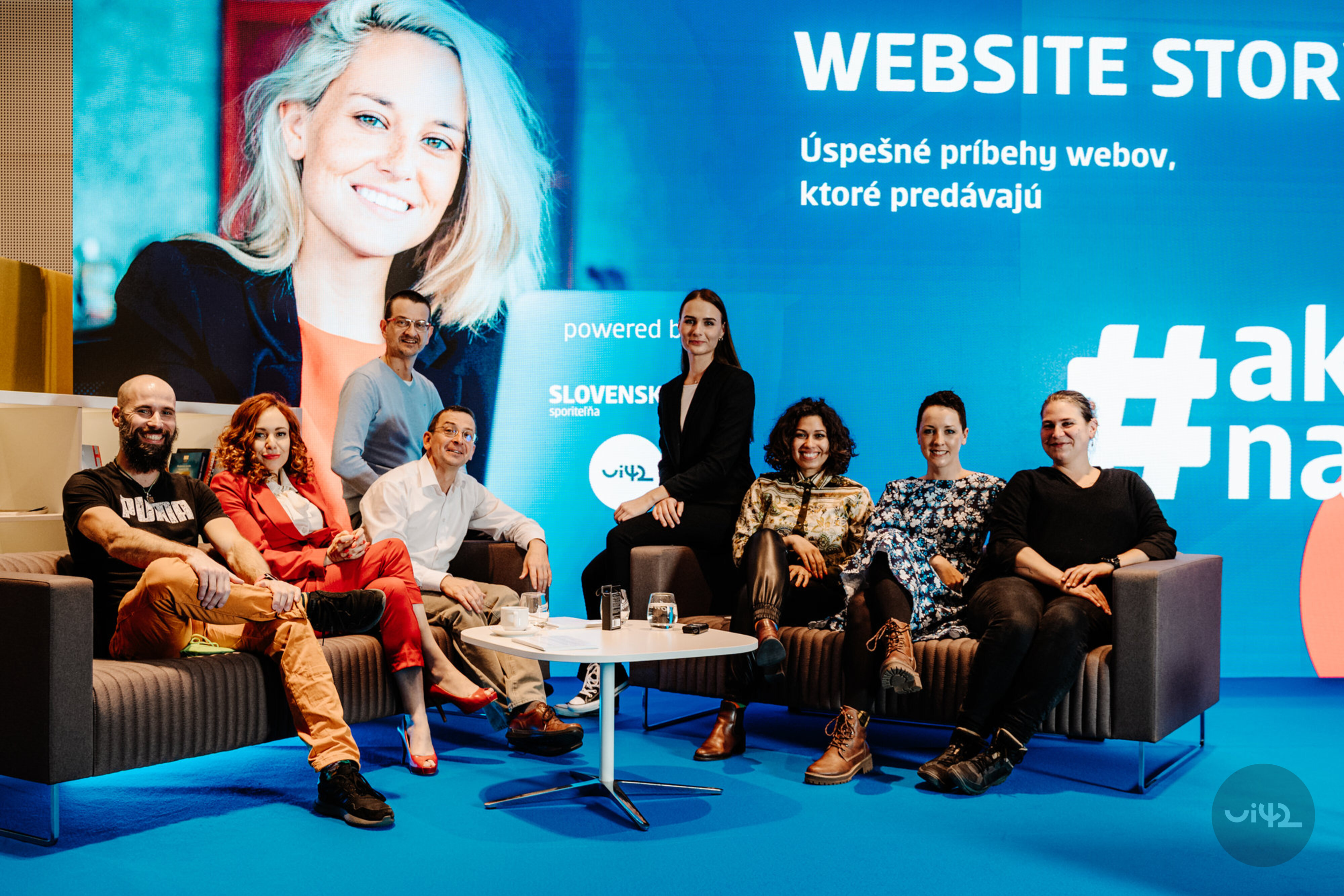Skupina lidí sedících na pohovkách během akce Website Story, s velkým modrým pozadím a nápisy o úspěšných příbězích webů.