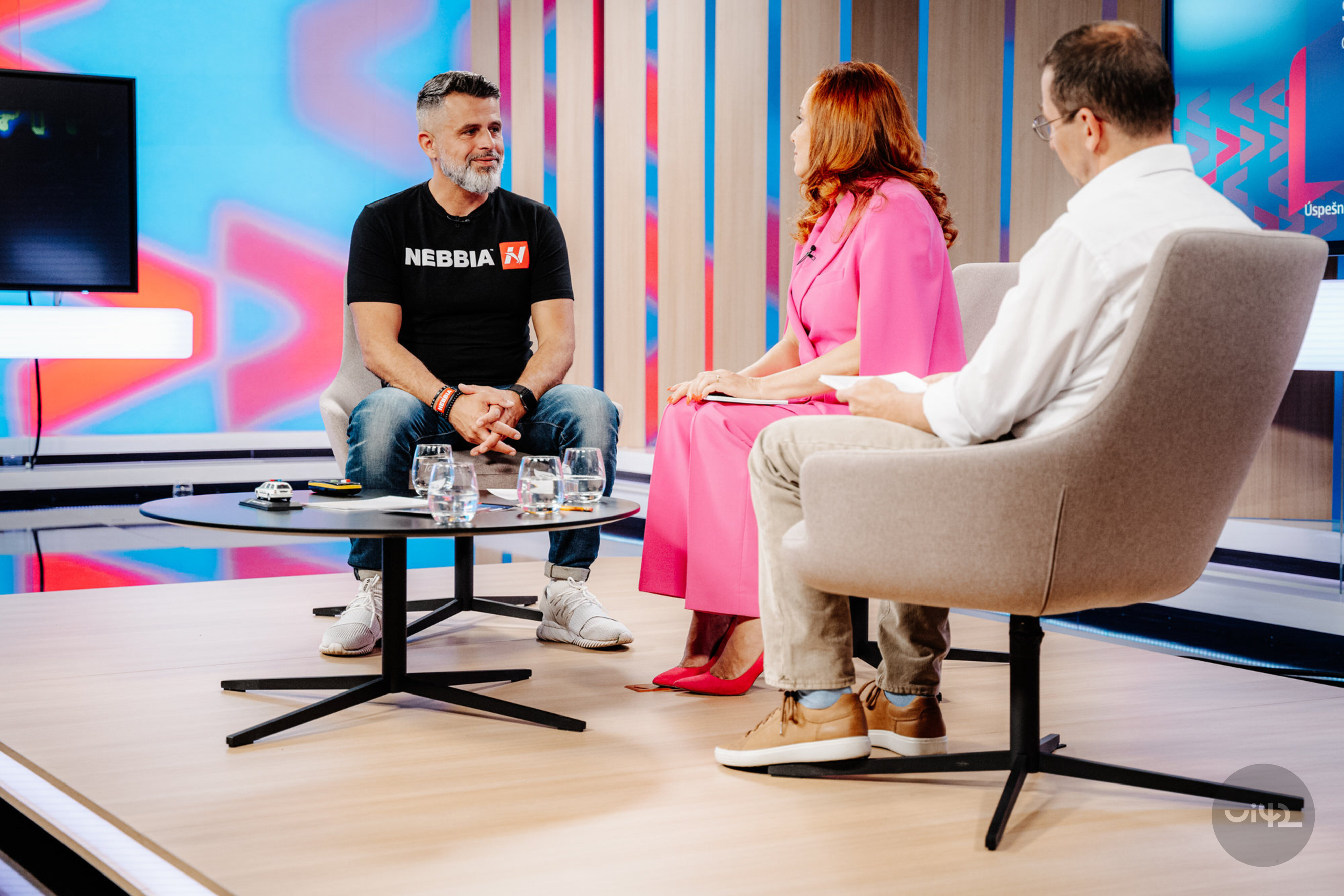 Diskuse tří osob v televizním studiu, muž v tričku Nebbia a žena v růžových šatech
