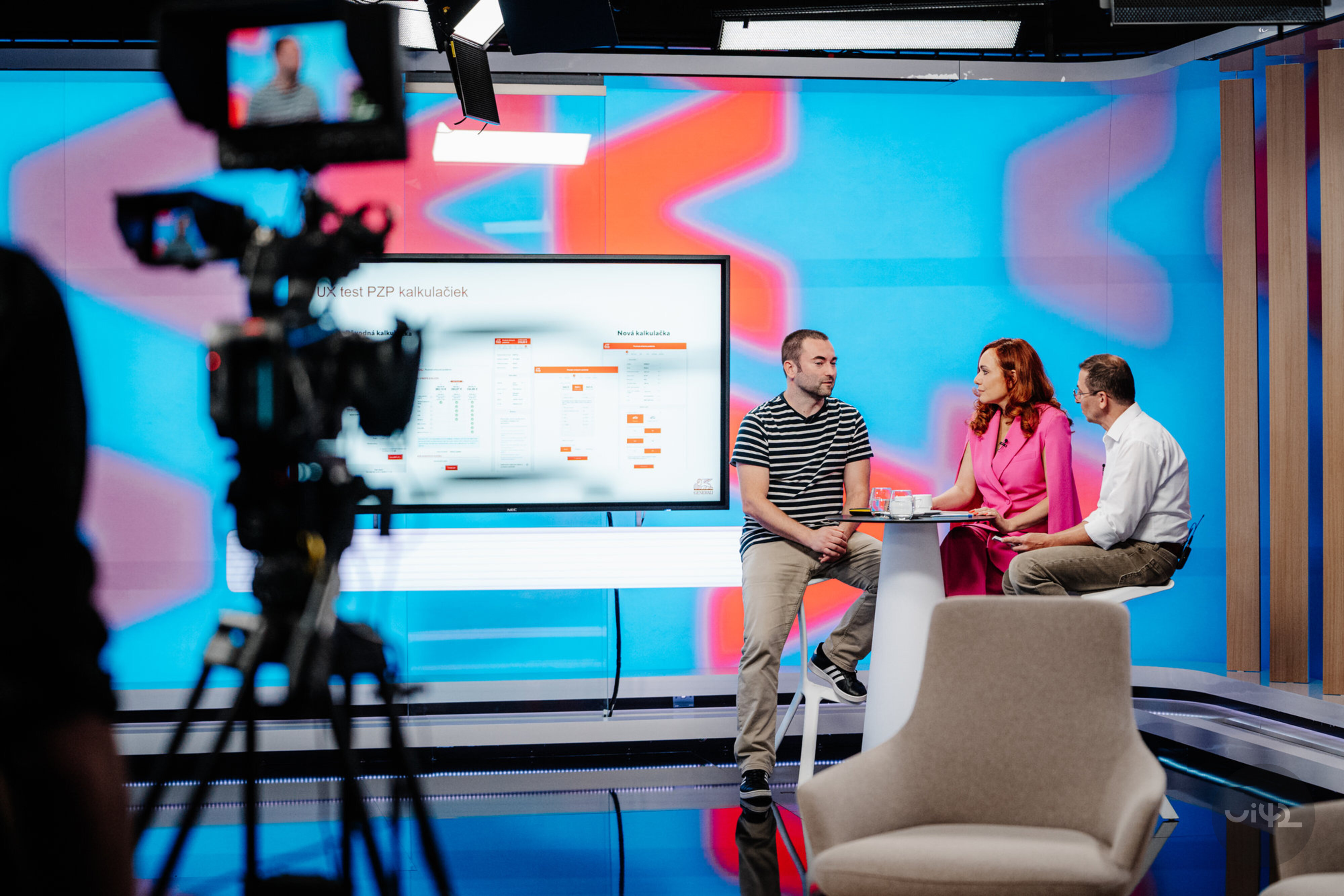 Televizní studio se třemi lidmi diskutujícími před obrazovkou, na které je zobrazen test kalkulaček. V pozadí je barevná obrazovka a kameraman s kamerou.