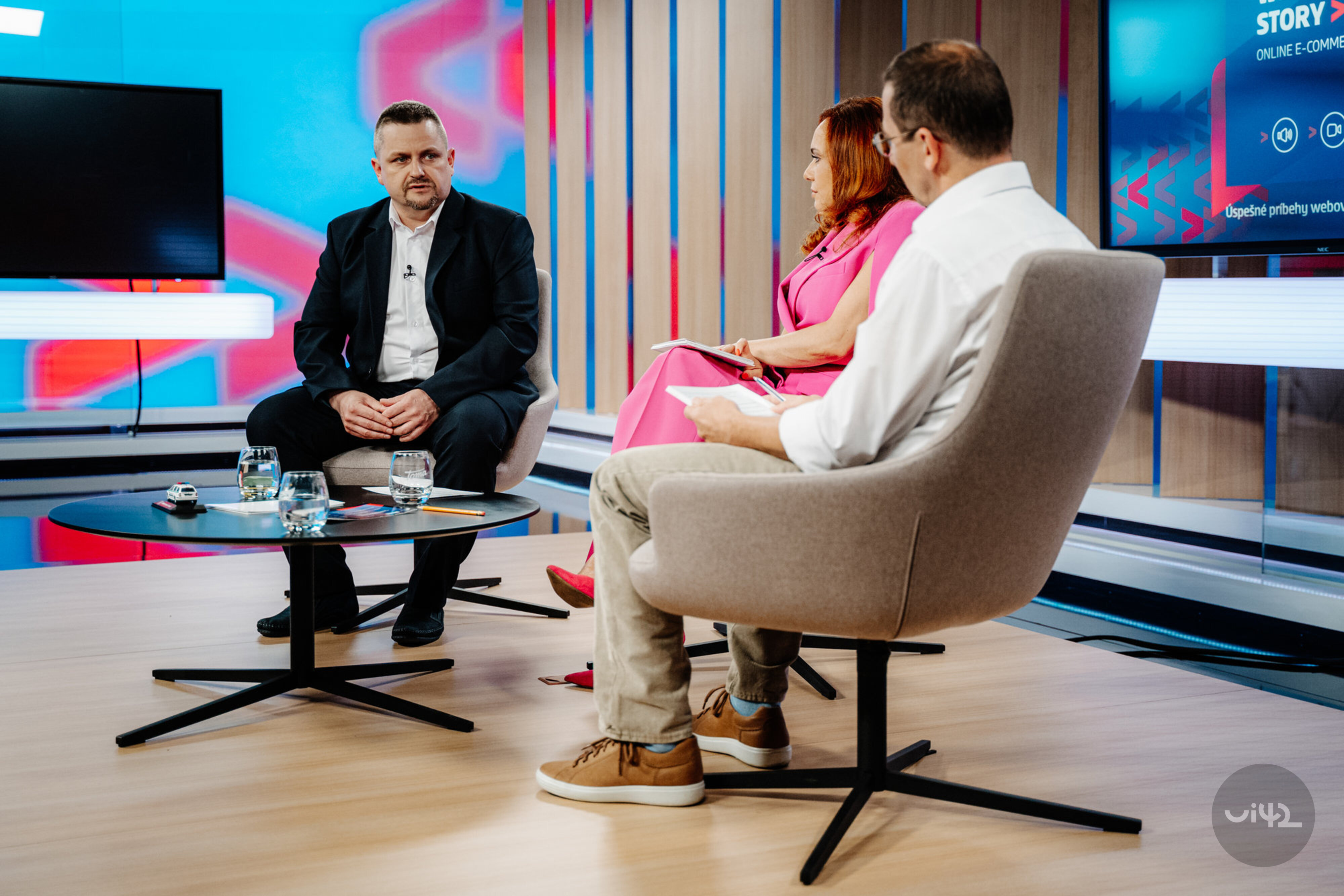Diskusní panel v televizním studiu se třemi lidmi sedícími v křeslech kolem stolku. Moderátor a hosté diskutují na téma přítomnosti nebo budoucnosti.