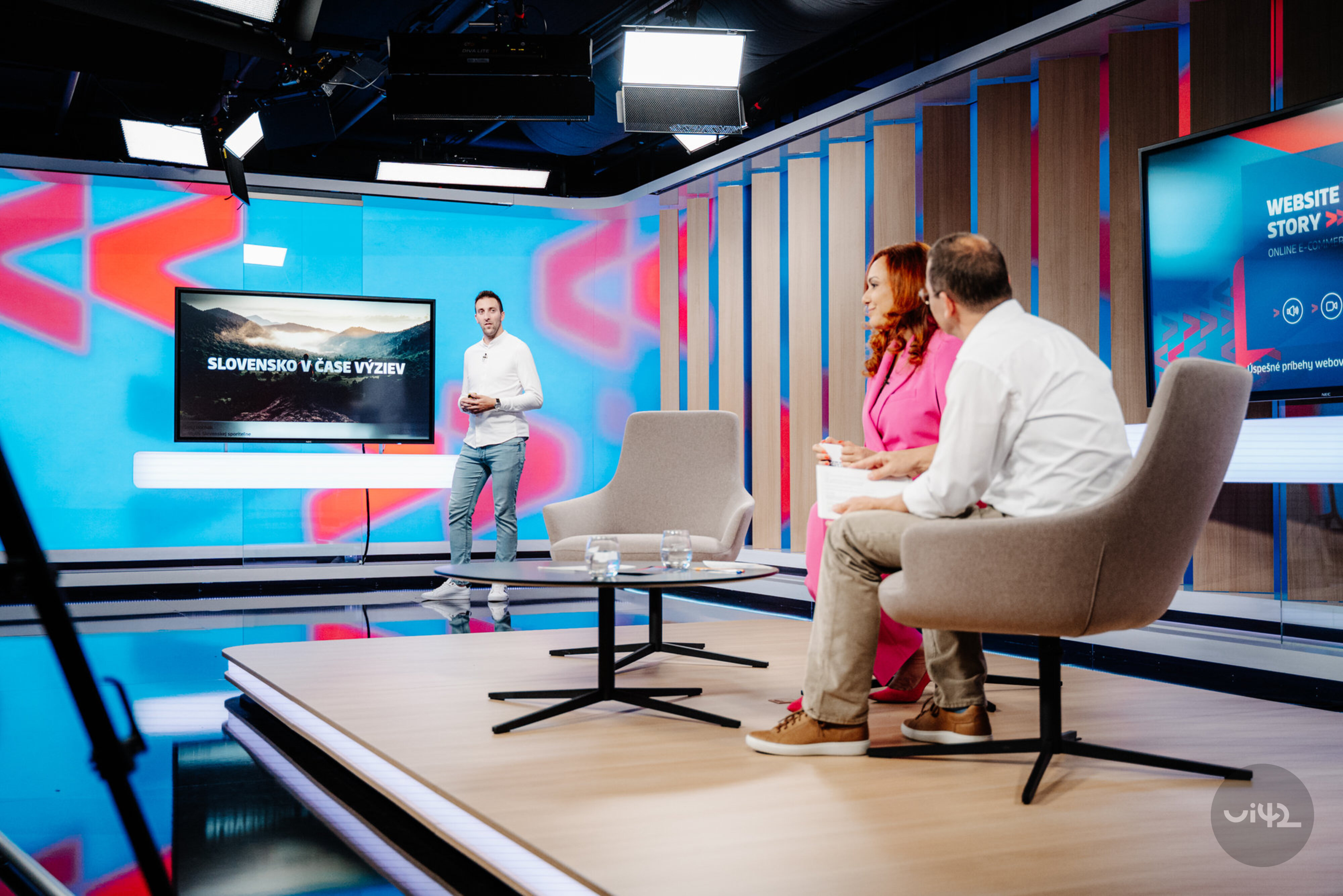 Diskusní studio se třemi lidmi, kde jeden stojí u obrazovky s textem 'Slovensko v čase výzev' a další dva sedí na židlích a sledují ho. Prostředí je moderní s barevnými světly a kamerou v popředí.