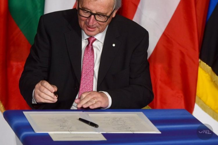 Muž podepisuje dokument na modrém stole s vlajkami v pozadí