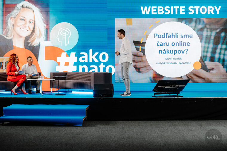 Online konference Website Story II. s prezentací na téma online nákupů. Na pódiu jsou dva moderátoři sedící na pohovce a jeden prezentující před velkým plátnem s nápisem 'Podlehli jsme kouzlu online nákupů?' a hashtagem #jaknat.