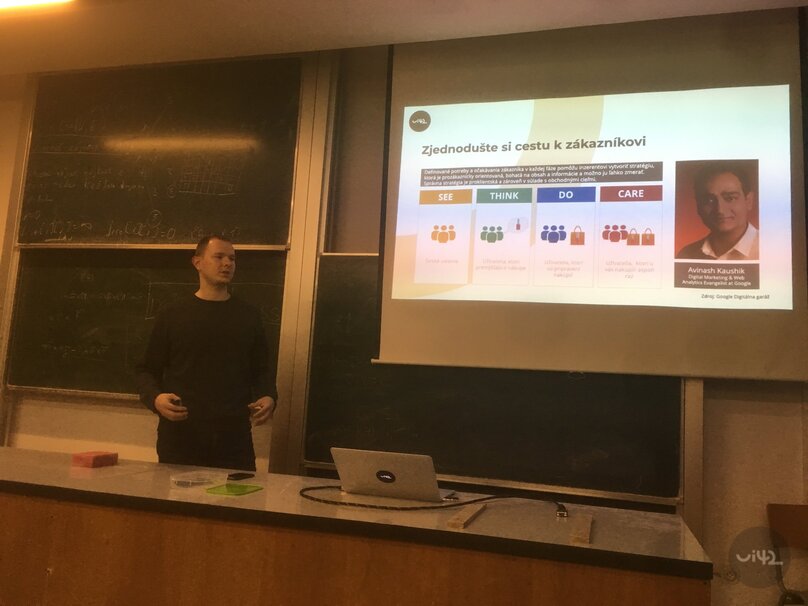 Prednáška na tému marketingovej stratégie v učebni s prezentáciou na plátne a tabuľami s poznámkami v pozadí.