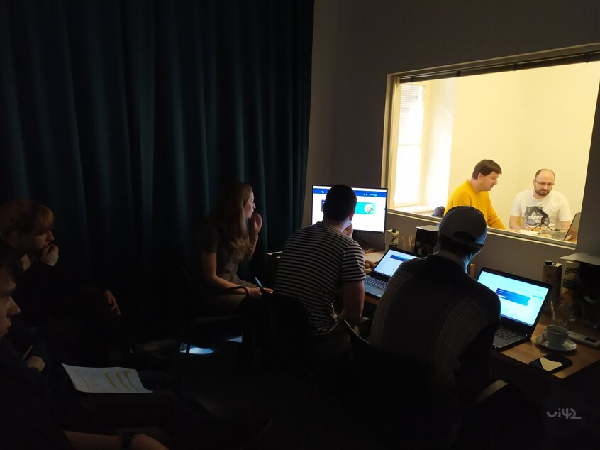 Skupina lidí pracujících v tmavé místnosti s počítači, dva muži za sklem v jiné místnosti.