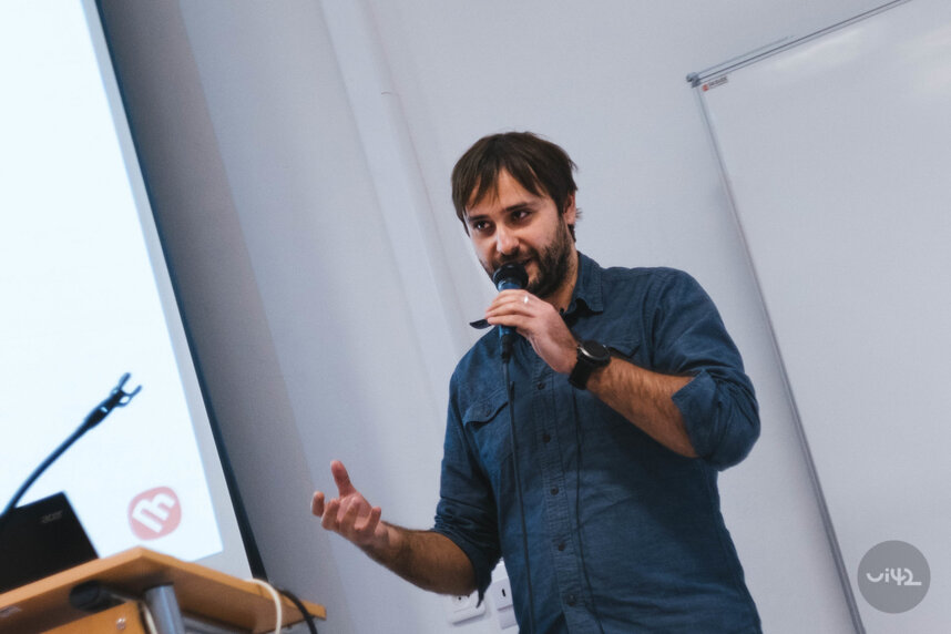 Muž mluví do mikrofonu na konferenci, oblečený v modré košili, stojí před projektorem a bílou tabulí.