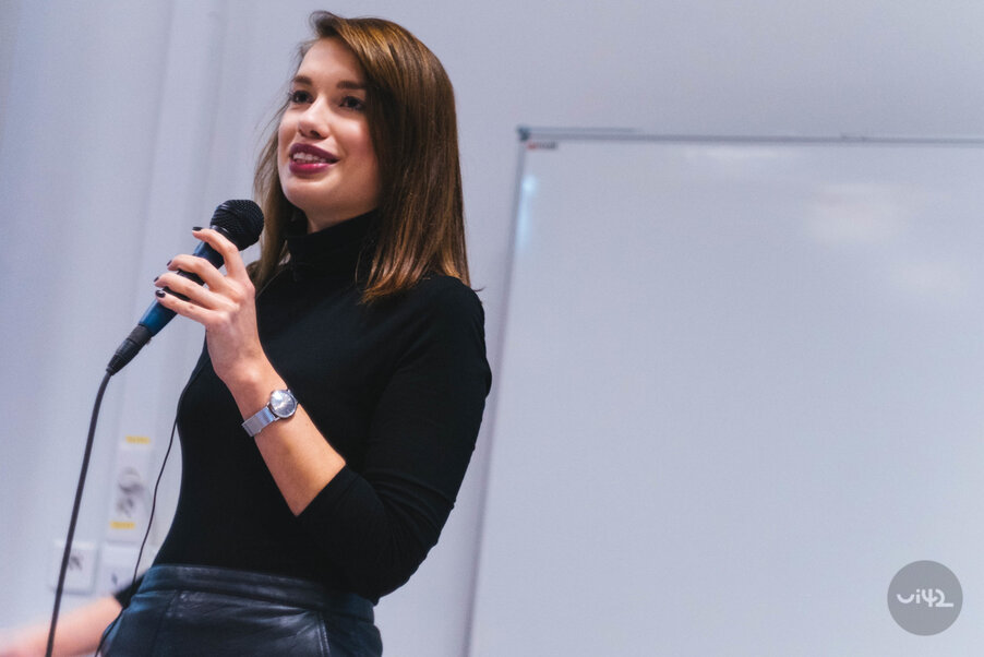 Žena drží mikrofon a mluví na konferenci před tabulí.