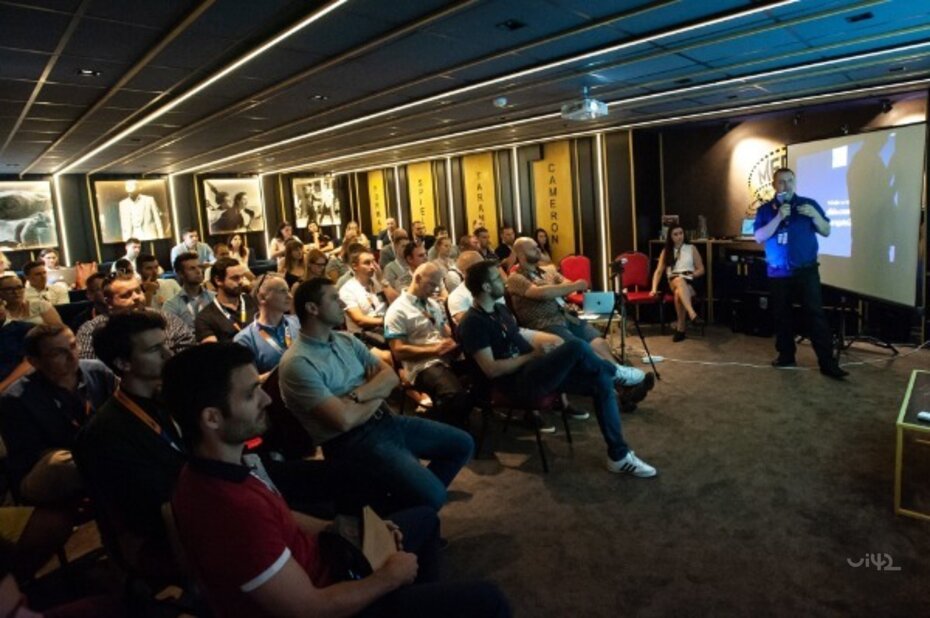 Účastníci konference MastersGate poslouchají prezentaci v tmavé místnosti.