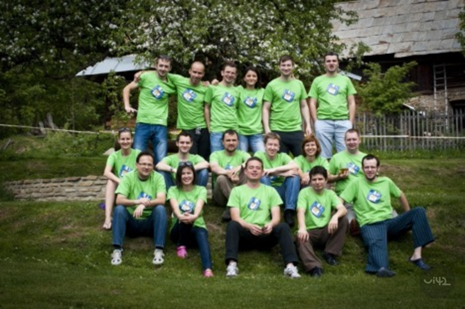 Skupina lidí v zelených tričkách sedí na trávě před chatou.