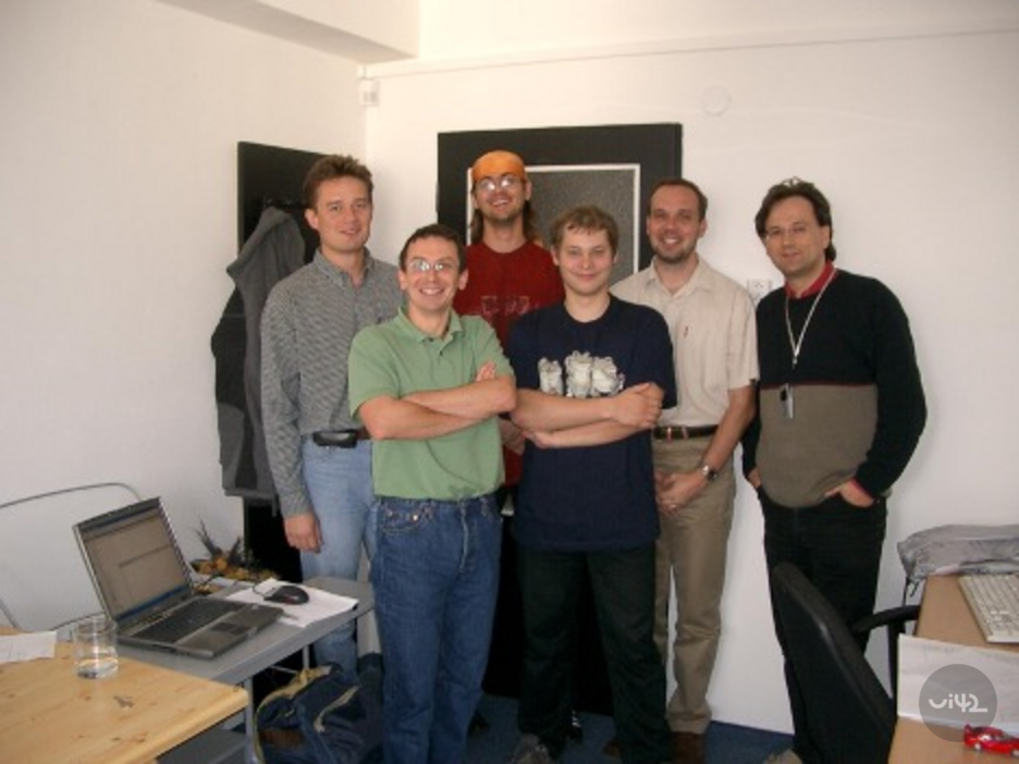 Skupina šesti mužů stojí spolu v kanceláři, usmívají se a jsou neformálně oblečeni. Na stole vedle nich je laptop a sklenice s vodou.