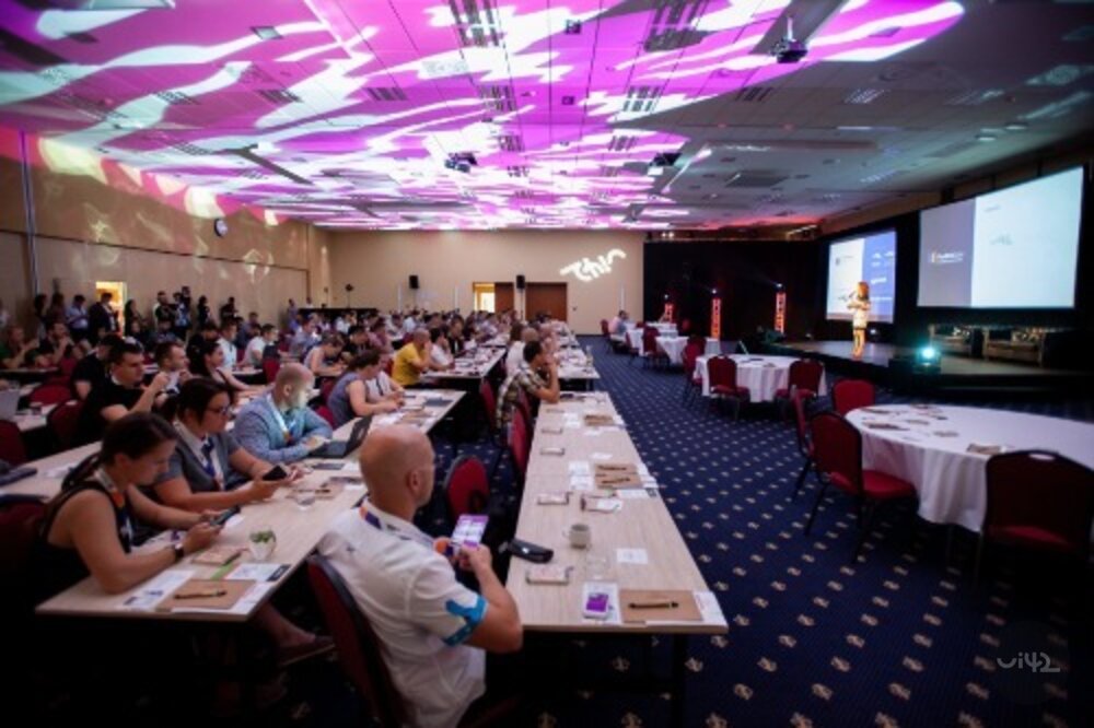 Účastníci na konferenci MastersGate sedí v konferenční místnosti, se světelnými efekty na stropě.