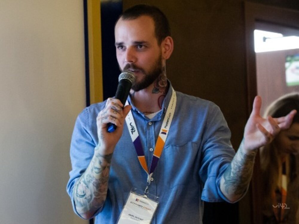 Muž s tetováním na rukou drží mikrofon a mluví na konferenci MastersGate.