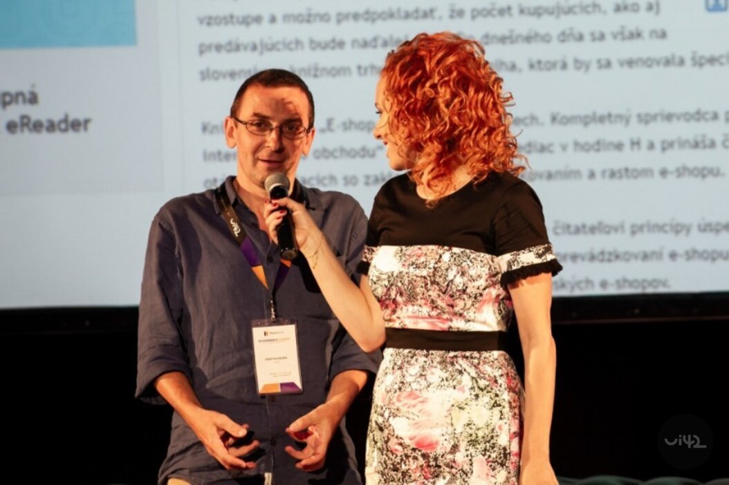 Dvě osoby na pódiu, muž s mikrofonem a žena v květinových šatech, prezentace na pozadí.