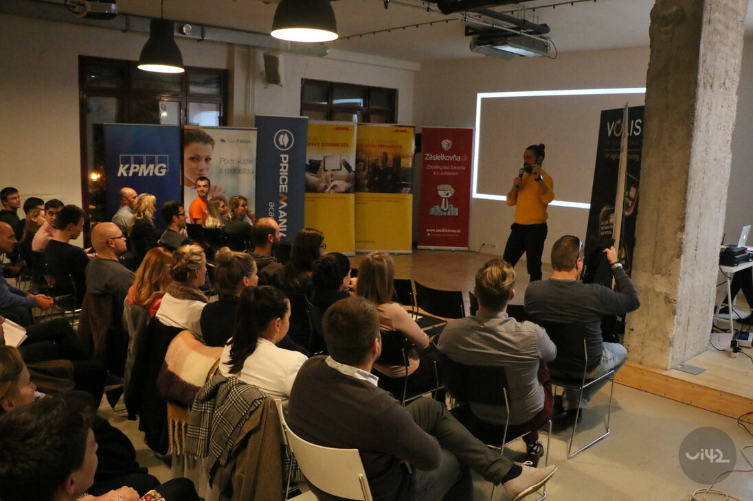 Přednáška na akci s účastí více lidí sedících na židlích, s reklamními bannery v pozadí.