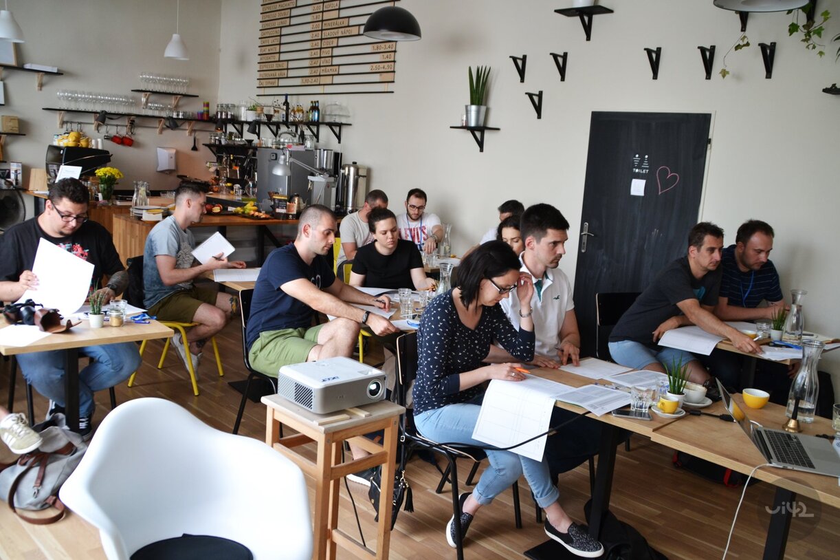 Účastníci na UX Bootcampu 2017 sedící v kavárně a soustředěně pracující na úkolech s projektorem na stole.