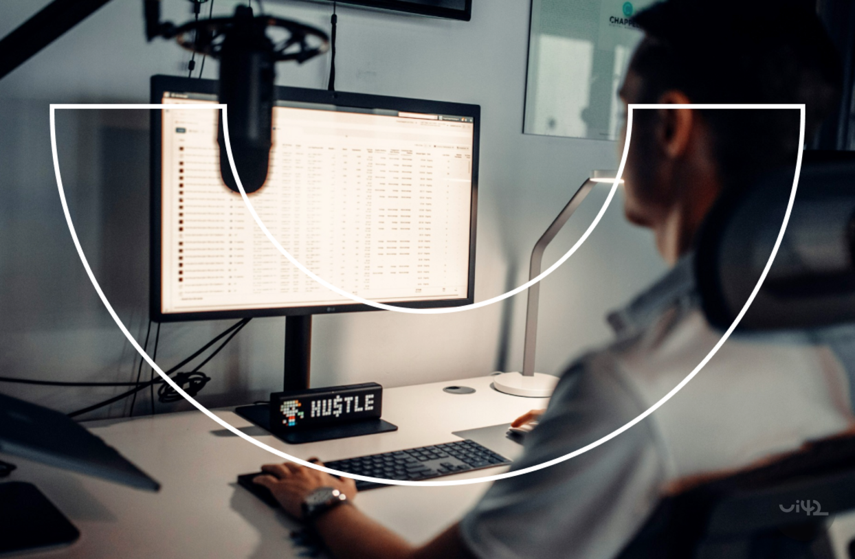 Osoba pracuje u počítače s velkým monitorem, na kterém jsou zobrazeny tabulky. Na stole je nápis 'HUSTLE'.