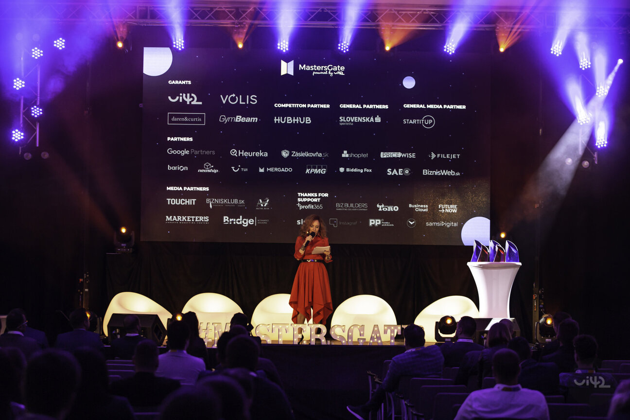 Ceremoniál MastersGate Awards 2019 s moderátorkou na pódiu a velkou obrazovkou zobrazující loga partnerů.