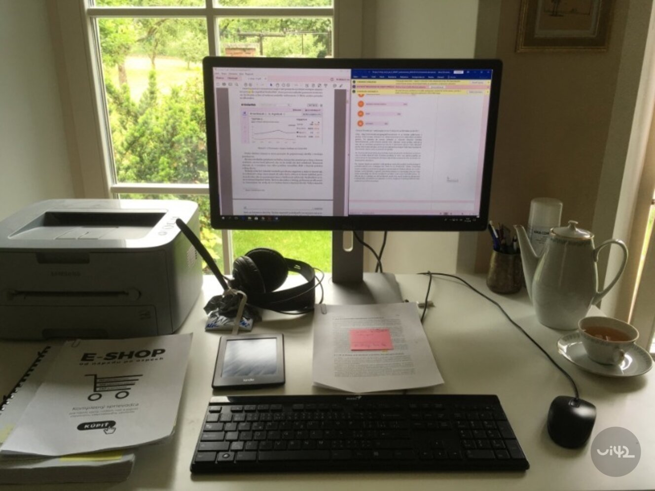Pracovní stůl s počítačovým monitorem zobrazujícím dokument, klávesnicí, myší, tiskárnou, sluchátky, knihou o e-shopech, čtečkou Kindle a šálkem čaje.