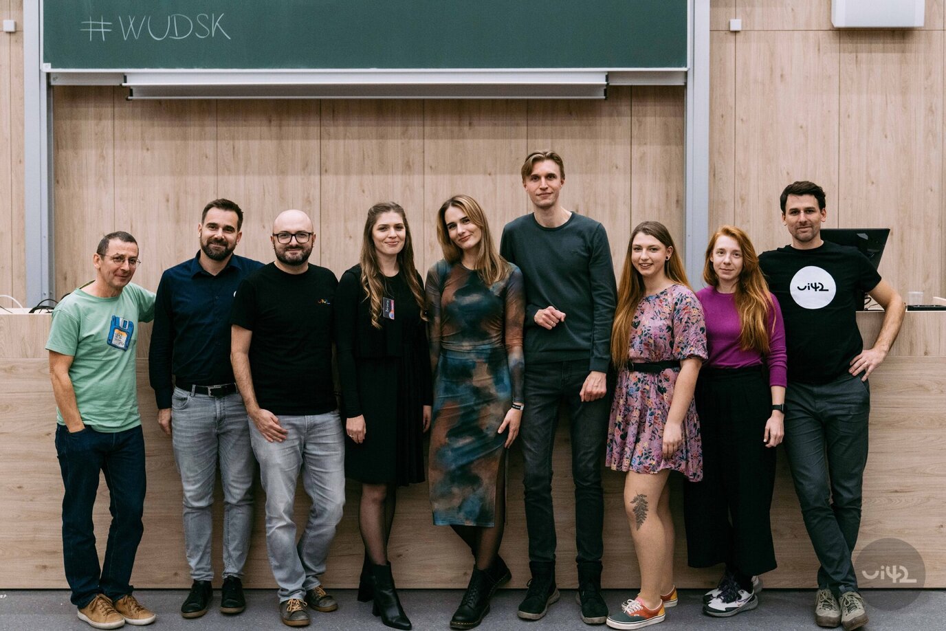 Skupina lidí pózujících před tabulí s nápisem #WUDSK, pravděpodobně na události World Usability Day.