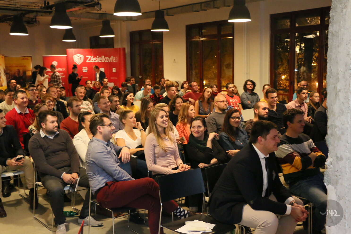 Skupina lidí na konferenci nebo semináři, někteří se usmívají a všichni pozorně sledují prezentaci. V pozadí jsou bannery s logem Zásilkovna.