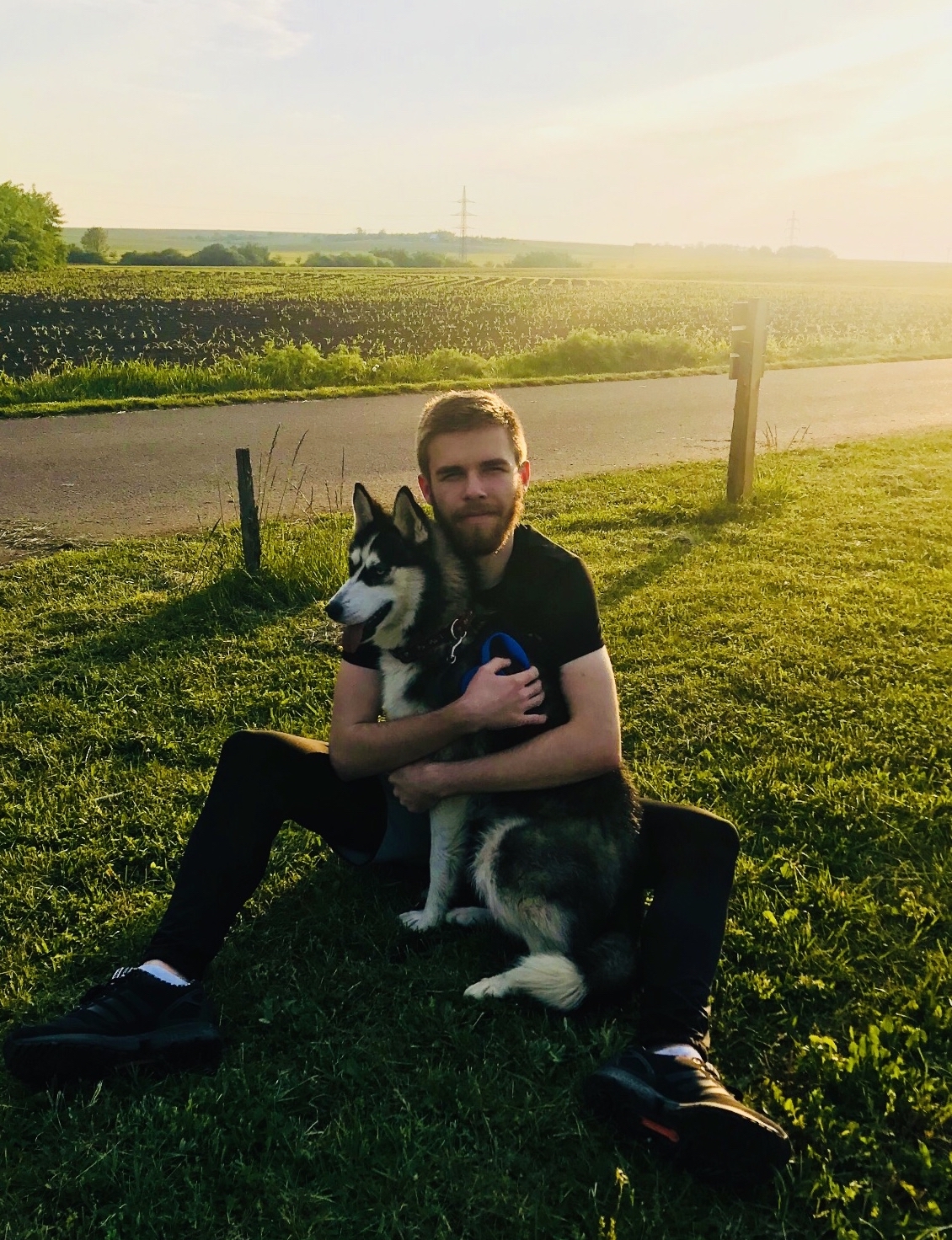 Muž sedící na trávě s huskym v přírodě při západu slunce.